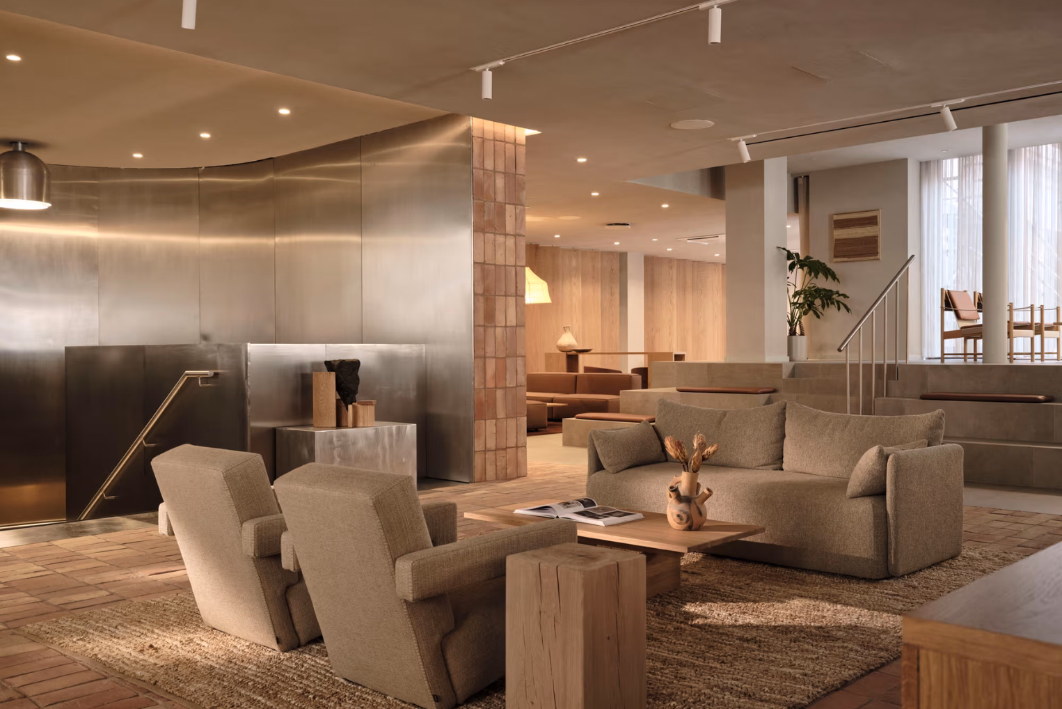 Armchairs and a sofa in shades of tan and beige on a rug around a wood table in a room with a silver metal wall and panelled flooring.