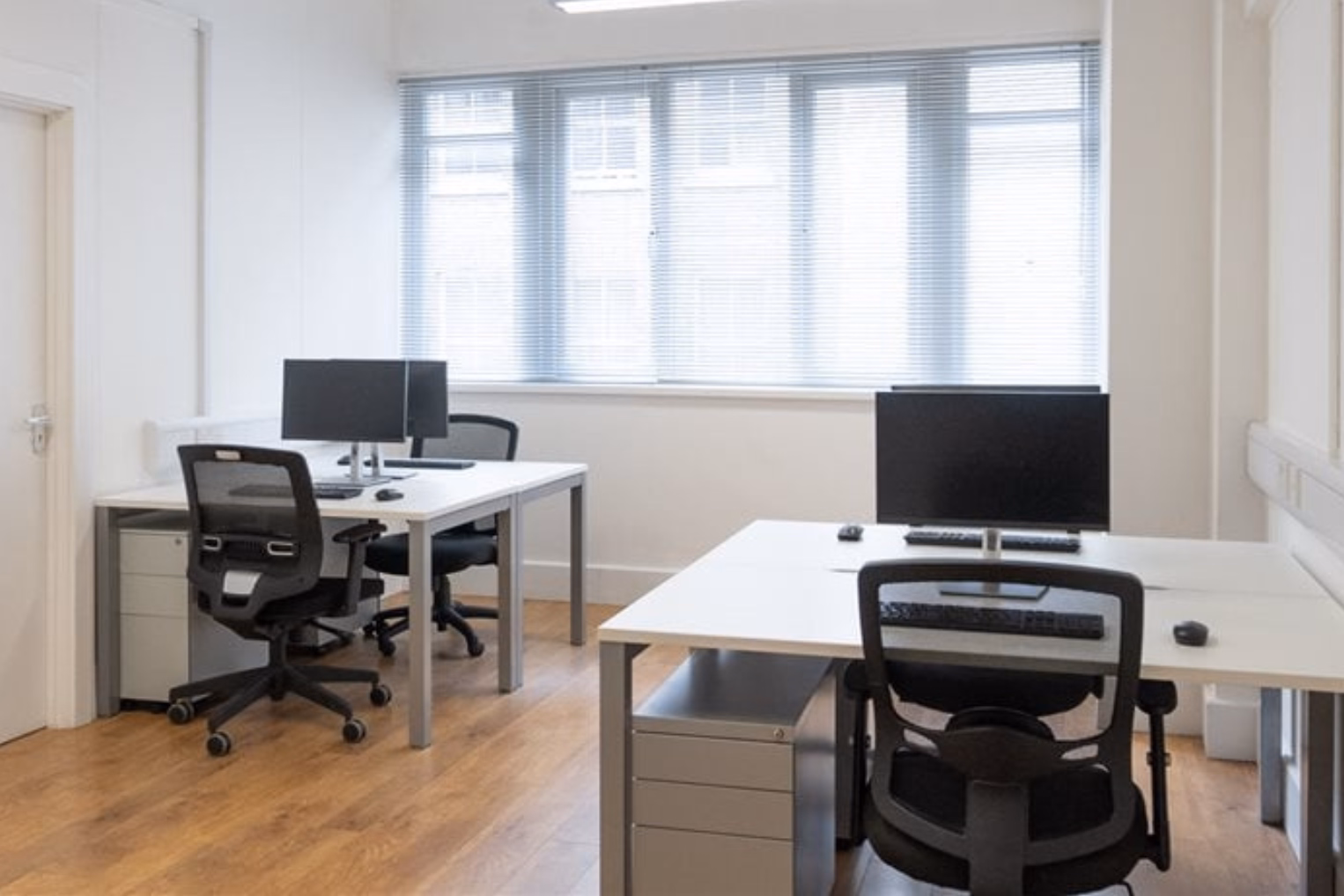 White desks with black chairs, black monitors, and grey filing cabinets sit on a wood floor in a room with a window and blinds.