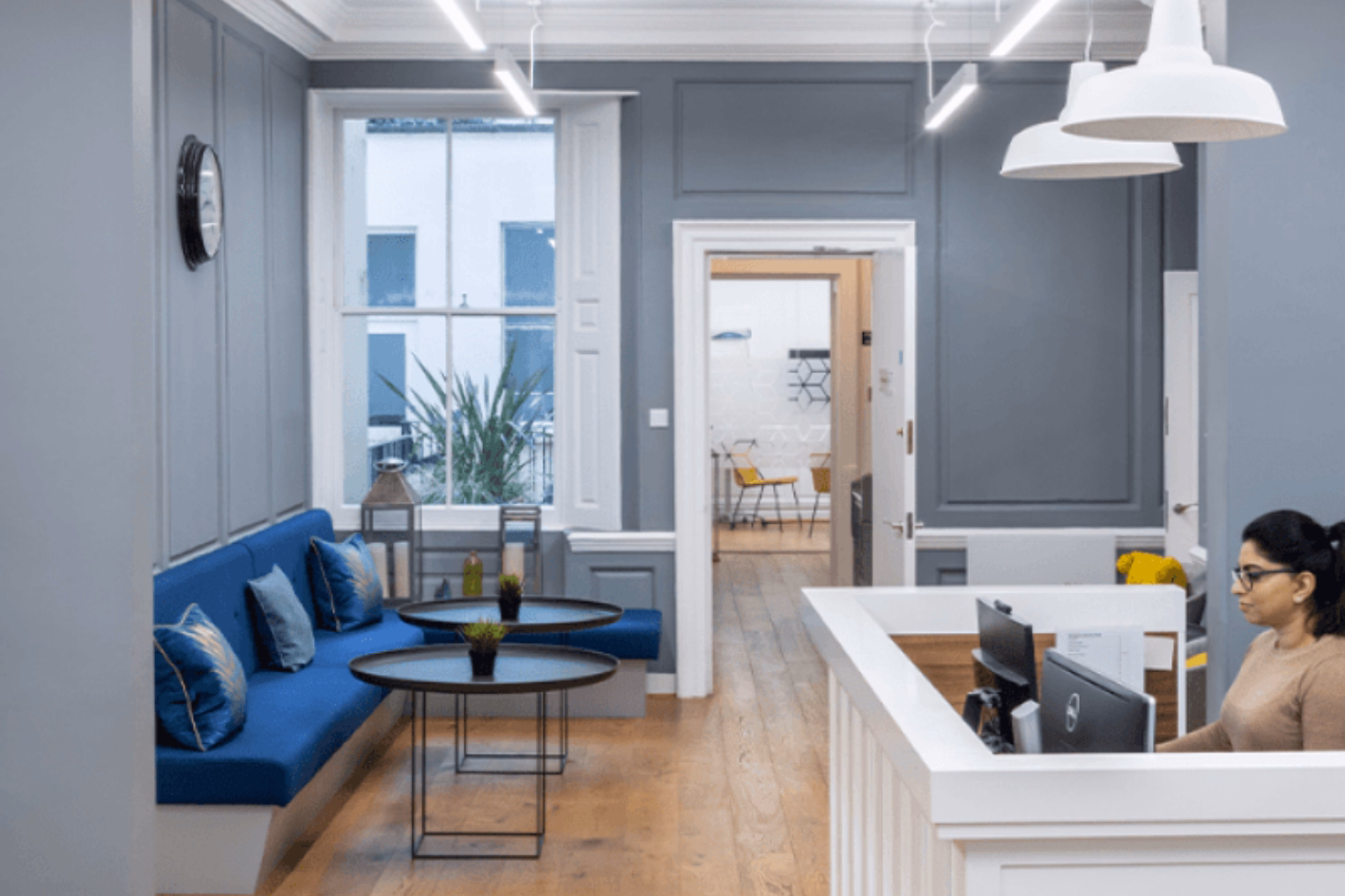 A person sits at a white desk and looks at a computer monitor. A blue bench with pillows and two black tables sit on a wood floor near a window.