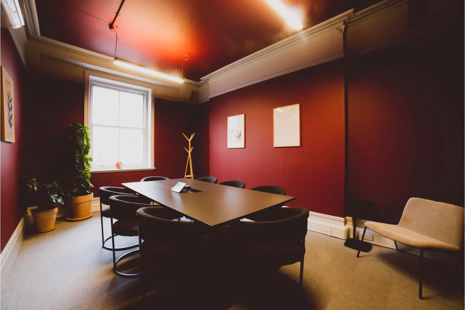 A brown table with black chairs sits in a room with red walls. A tan chair stands in the corner near a window and plants in tan pots on a grey carpet.