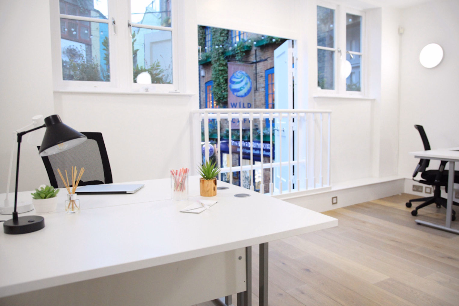 White desks with black chairs and a black lamp sit on a wood floor in a room with white walls. An open door with a white railing overlooks a street with buildings.