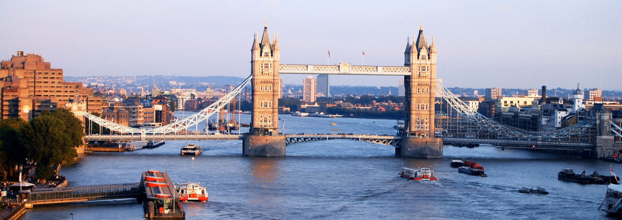 White and red boats travel on the River Thames toward Tower Bridge and city buildings. People stand on a pier and on the boats under a light sky.