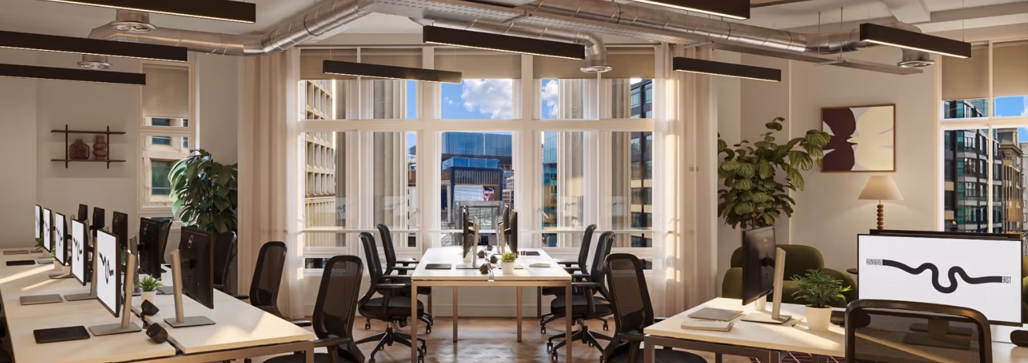 Light wood desks with black chairs and monitors on a wood floor in an office with windows and plants. Silver pipes and black lights hang from the ceiling near a green chair.