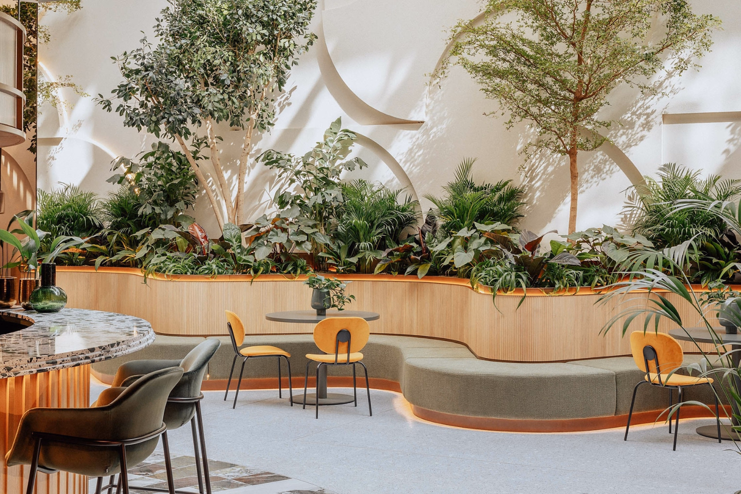 Yellow chairs and grey tables near a curved green bench and indoor plants. A speckled table and green chairs sit on a grey floor near a white wall with circular patterns.