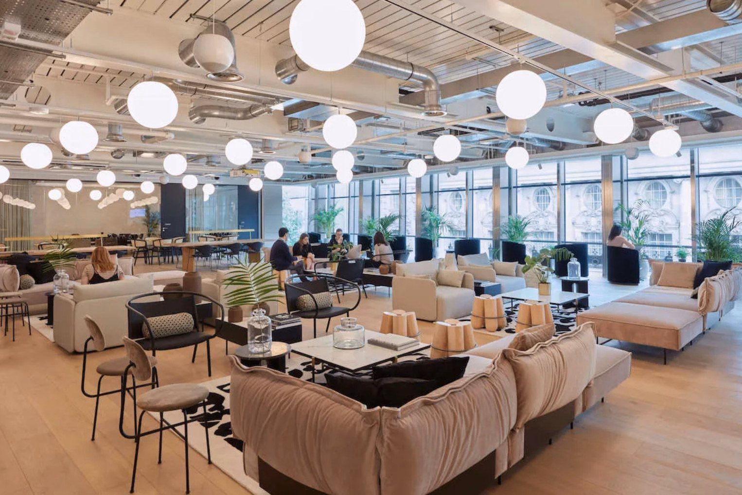 People sit on cream sofas and black chairs talking in a room with white tables, sphere lights, plants, and the furniture is on wood floors near floor-to-ceiling windows.