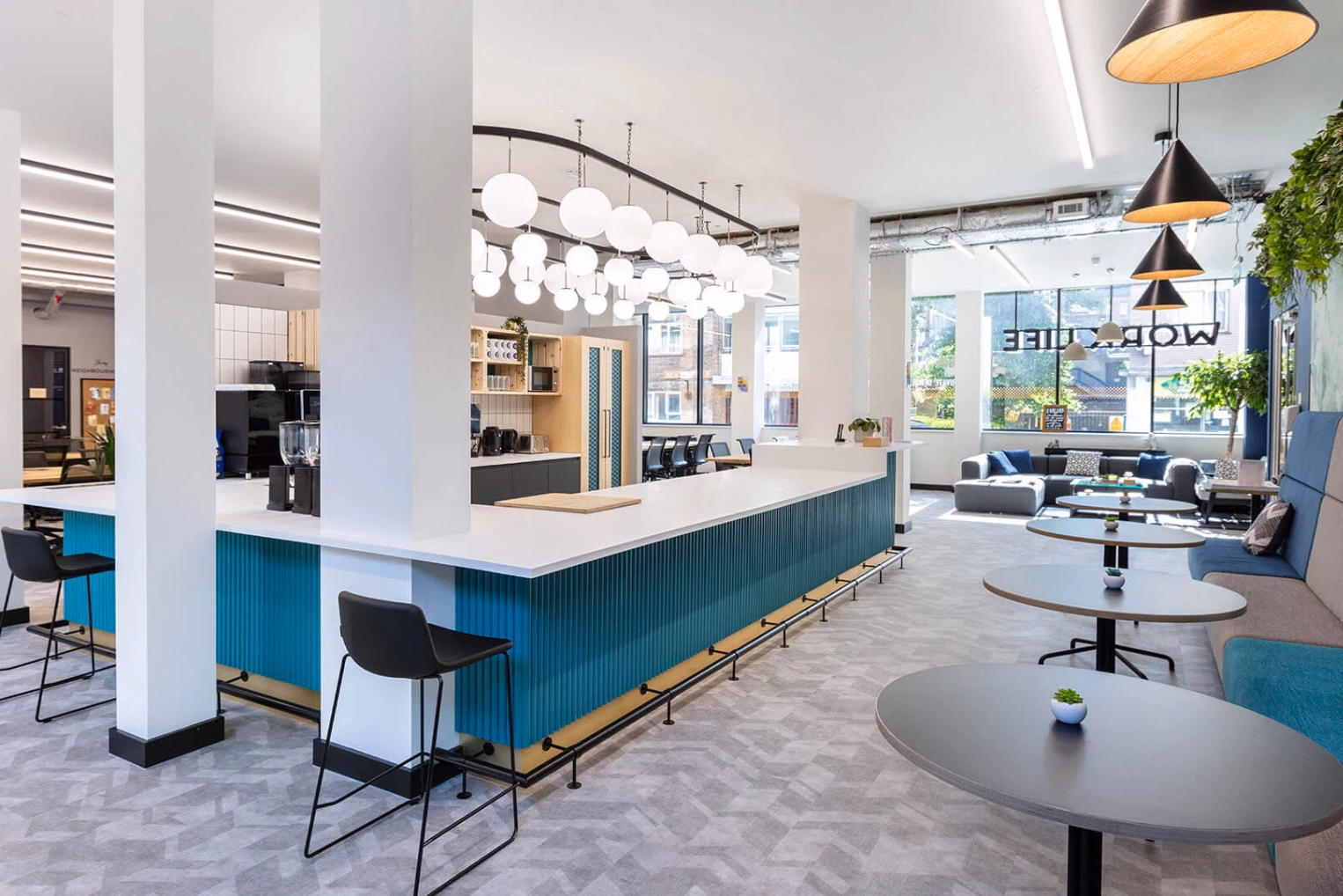 A teal and white counter with black stools under globe lights near round grey tables and a blue bench. A set of grey sofas in the background near a window.