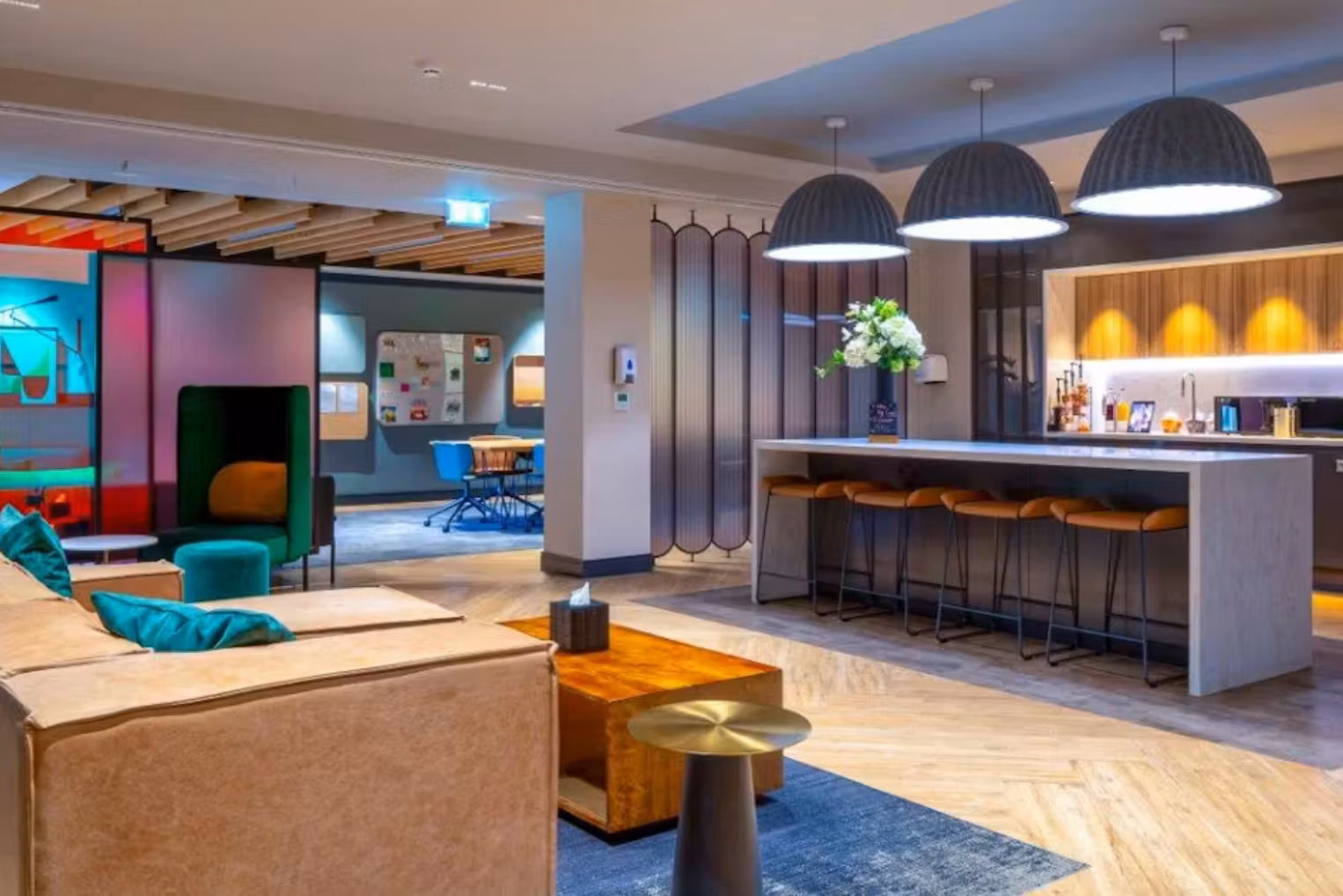 A cream sofa with teal pillows sits on a blue rug near a wood table. A grey counter with brown stools stands under black lights near a green chair, a teal ottoman, and a pink screen.