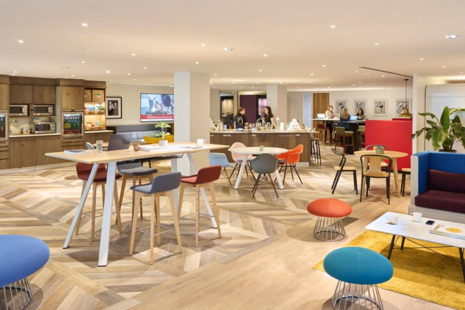 People talk and work at tables near wood kitchen cabinets. A high wood table with grey and red stools stands on a wood floor near blue, red, and teal stools, a blue booth, and a red screen.
