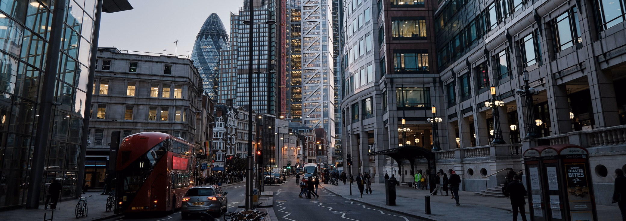 A red bus and silver car travel on a street while people cross the road and walk past red phone booths. Skyscrapers, including the Gherkin in the background under a grey sky.