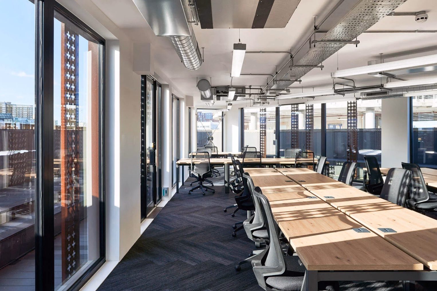 Rows of wood desks and black chairs on a grey carpet in a room with floor-to-ceiling windows and silver pipes on the ceiling.