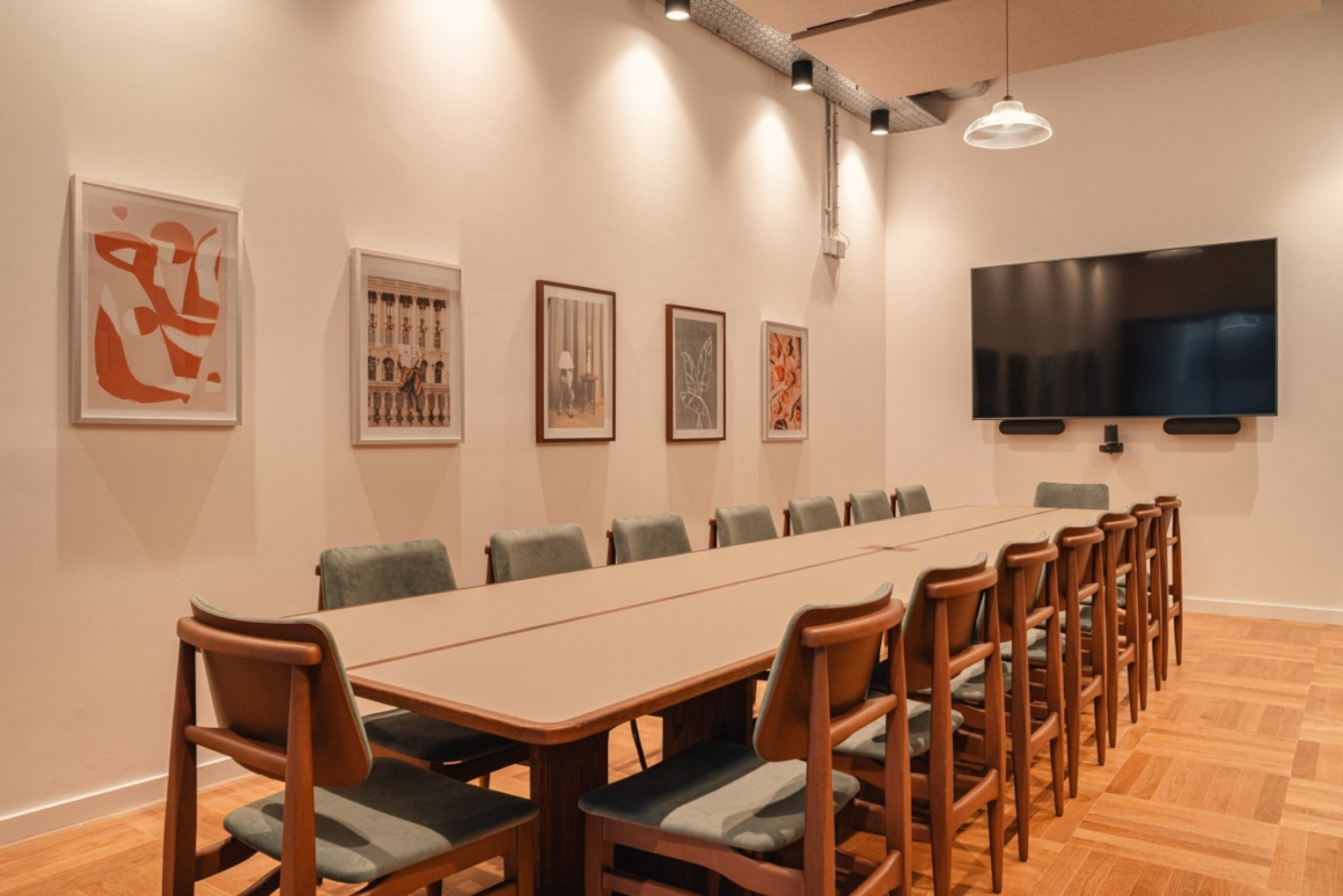 Green chairs with brown frames around a tan table on a wood floor. Pictures and a black screen hang on white walls under lights.