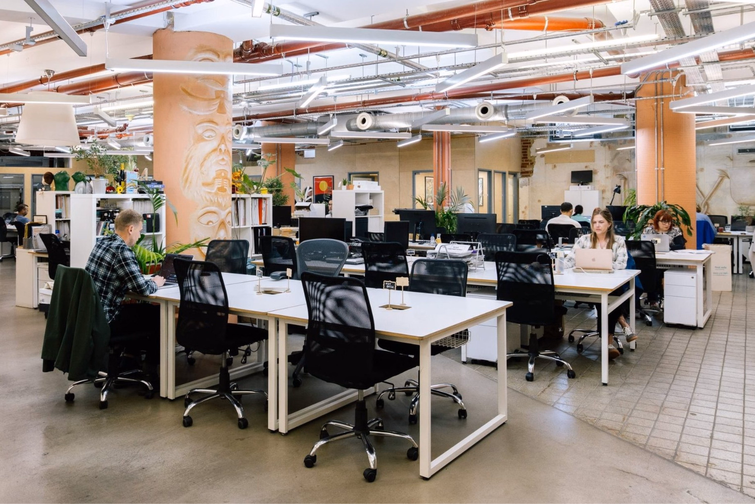 People sat at white desks with black chairs, working on computers in an office with white shelves. Red and silver pipes and white lights hang from the ceiling near tan pillars and plants.