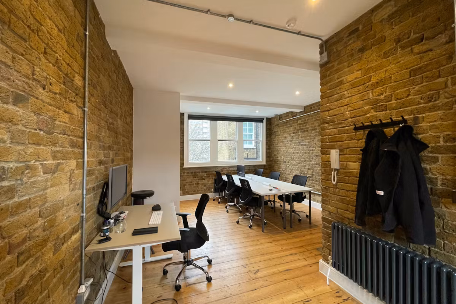 A wooden desk and white desks with black chairs on a wood floor between brick walls. A window, coats on a rack, and a radiator are in an office with pipes on the ceiling.