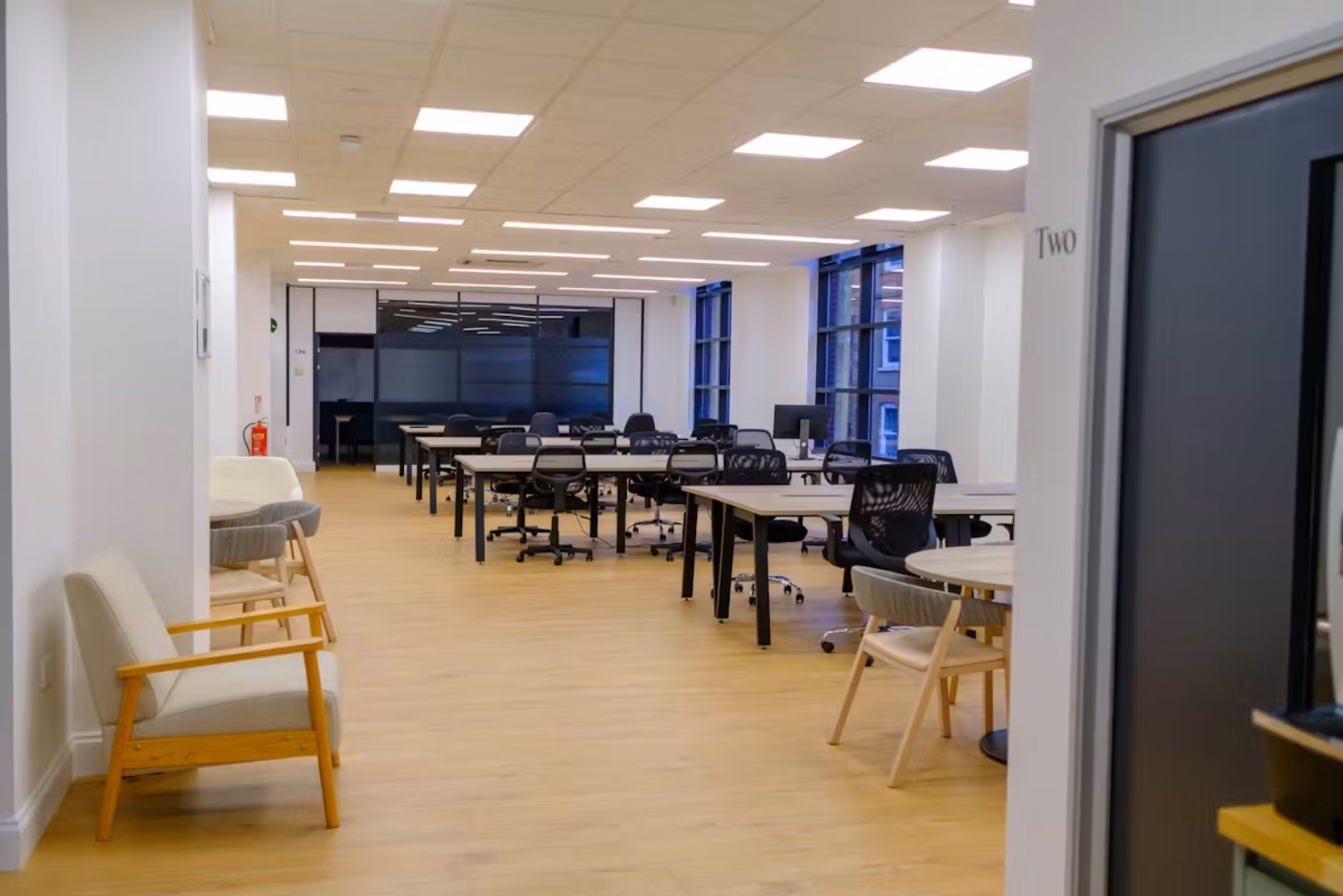 Light wood desks and black chairs on a wood floor near cream armchairs and grey chairs. Ceiling lights illuminate white walls and a grey door in an office.