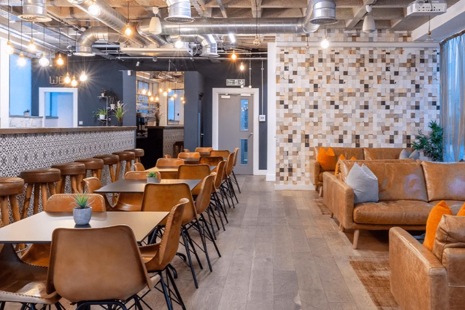 Brown chairs and black tables sit on a wood floor near brown leather sofas with orange and grey pillows. Brown stools line a counter under ceiling lights and silver pipes.