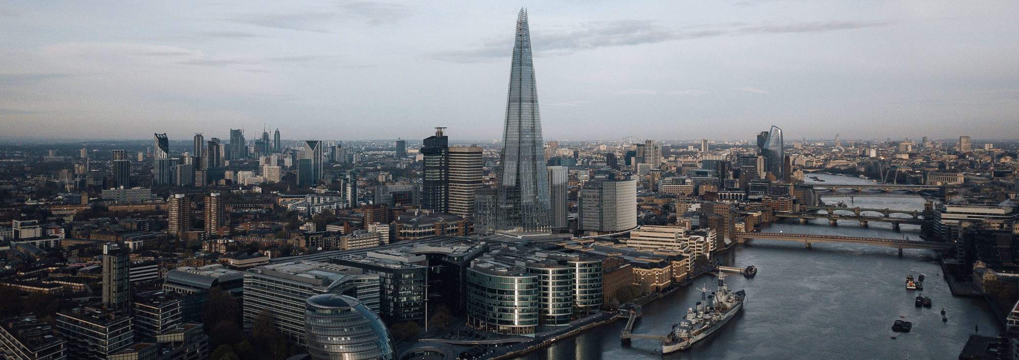 Buildings and skyscrapers, including the Shard, along a river with bridges and boats under a grey sky.