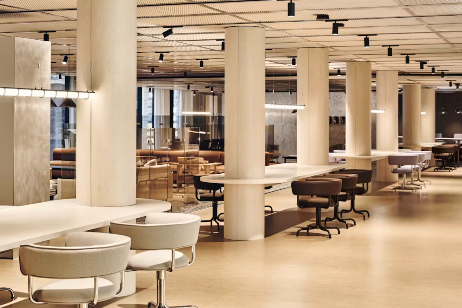 White tables with cream and brown chairs around cream pillars. Tan sofas and black lights on a ceiling are in a room.