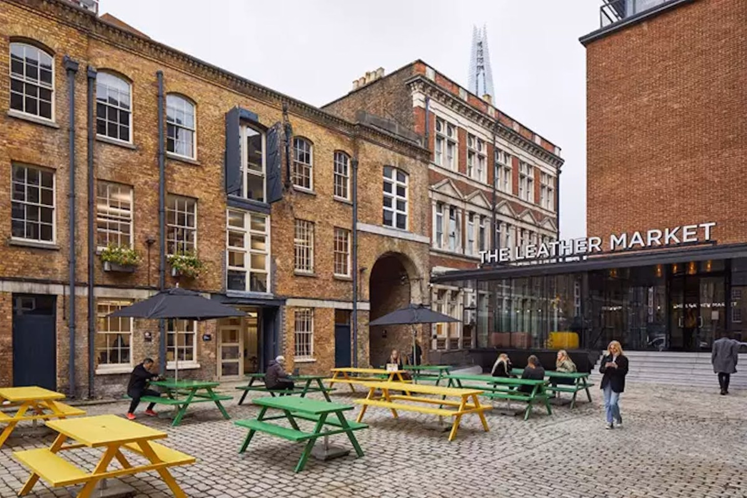  People sat at green and yellow tables in a courtyard while a person walks and looks at a phone. Brick buildings and a sign for "THE LEATHER MARKET" in the background under a grey sky.