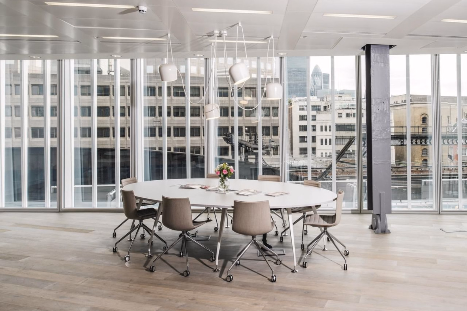 A white table and grey chairs sit on a wood floor under white lights. Windows show buildings and a skyscraper in the background.