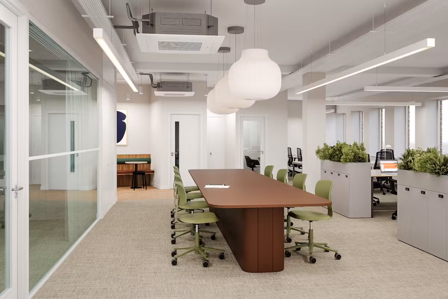 Green chairs sit around a brown table on a grey carpet. Grey cabinets with plants and desks in a room with white walls under big white lights.