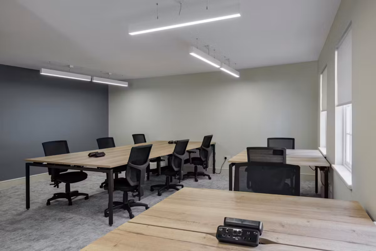 Wood desks and black chairs on a grey carpet in a room with white and grey walls. White lights hang from the ceiling near windows.
