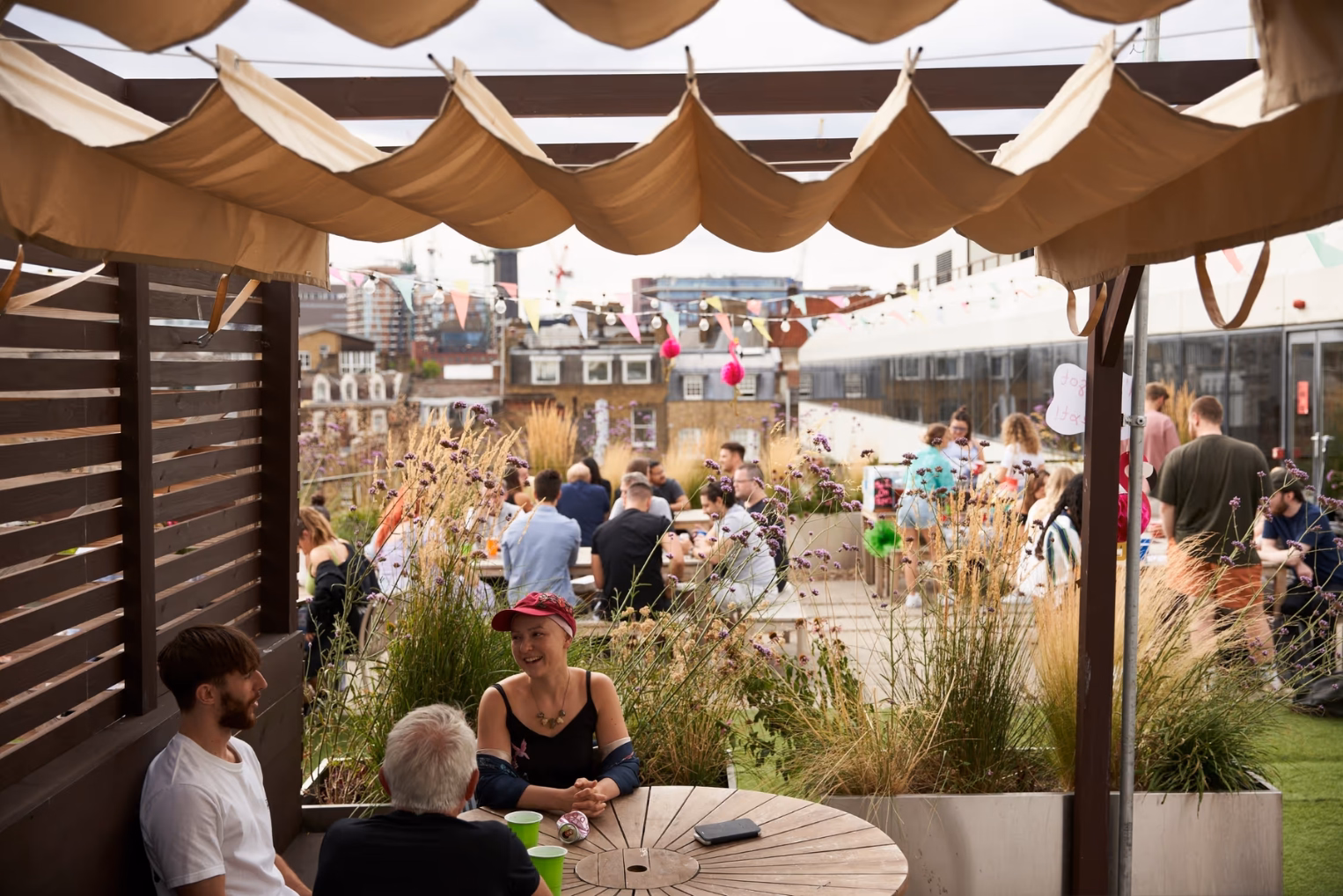A crowded rooftop terrace featuring people sitting at wooden tables under a beige fabric canopy, surrounded by tall grass and colourful bunting.