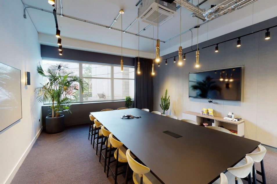 A long black table with chairs sits in a meeting room with pendant lights, a plant, and a television.