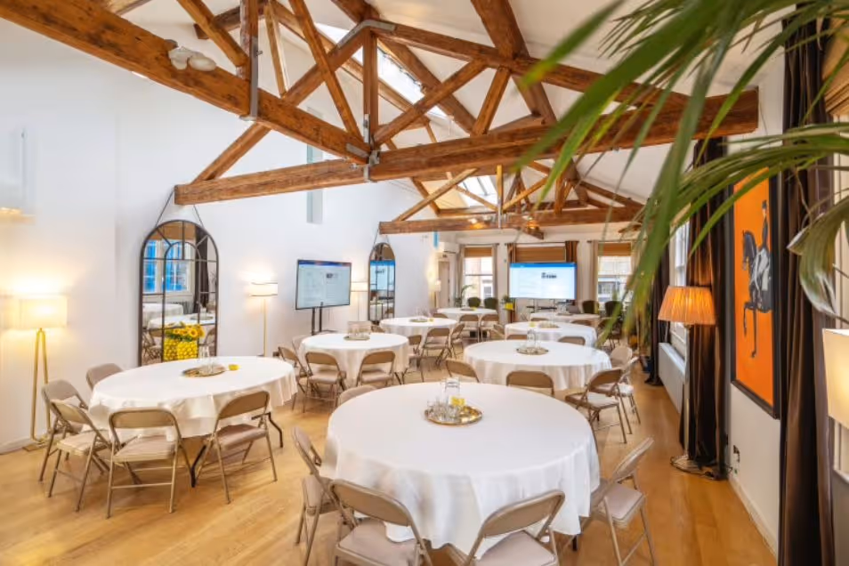 Round tables with white cloths are set up in a room with wooden ceiling beams and floor lamps.