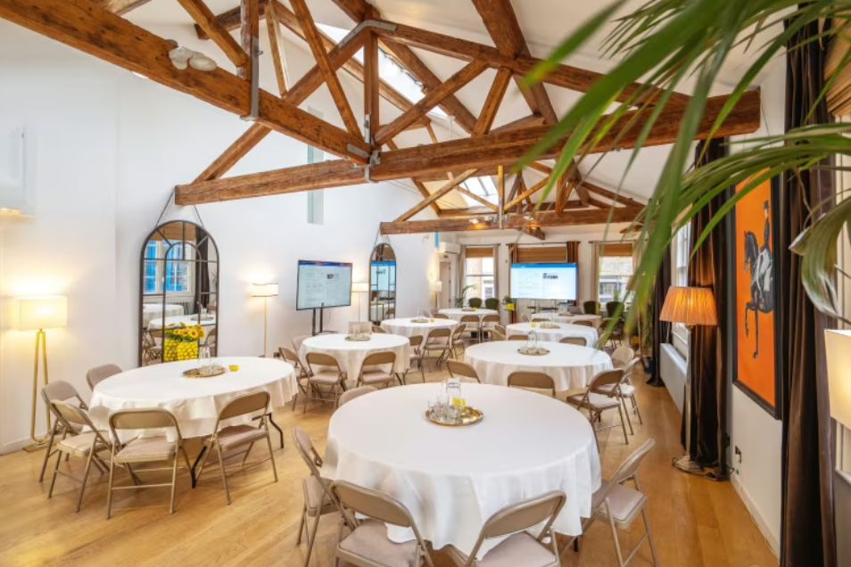 Round tables with white cloths are set up in a room with wooden ceiling beams and floor lamps.