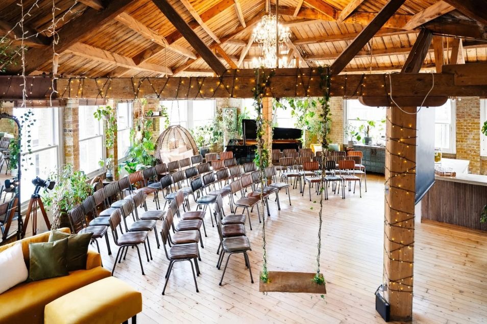 Rows of chairs are set up for an event in a room with wooden ceiling beams, a swing, and plants.