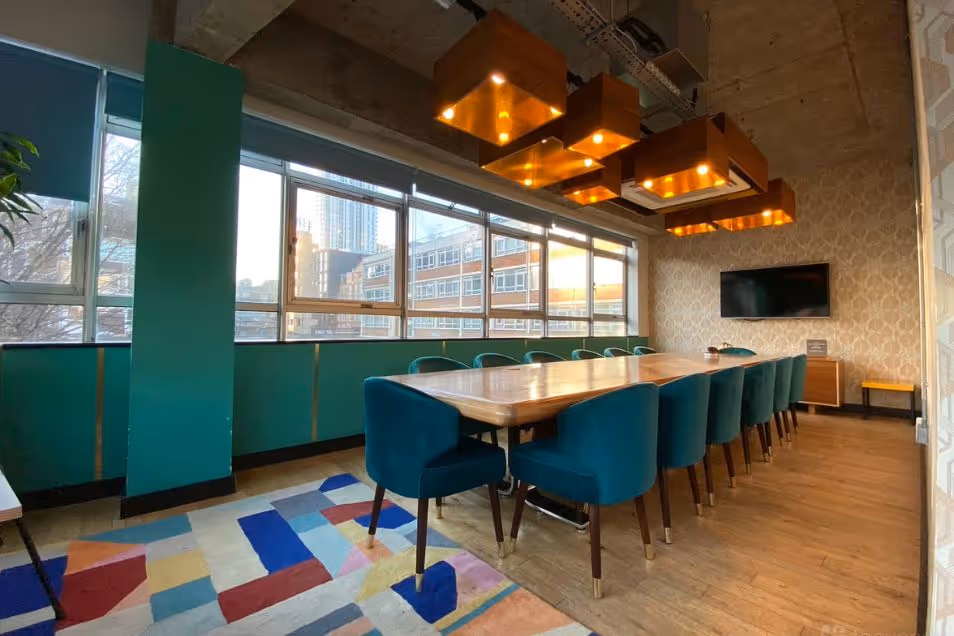 Long wooden conference table with teal chairs in an industrial-style room featuring copper box lighting and a geometric rug.