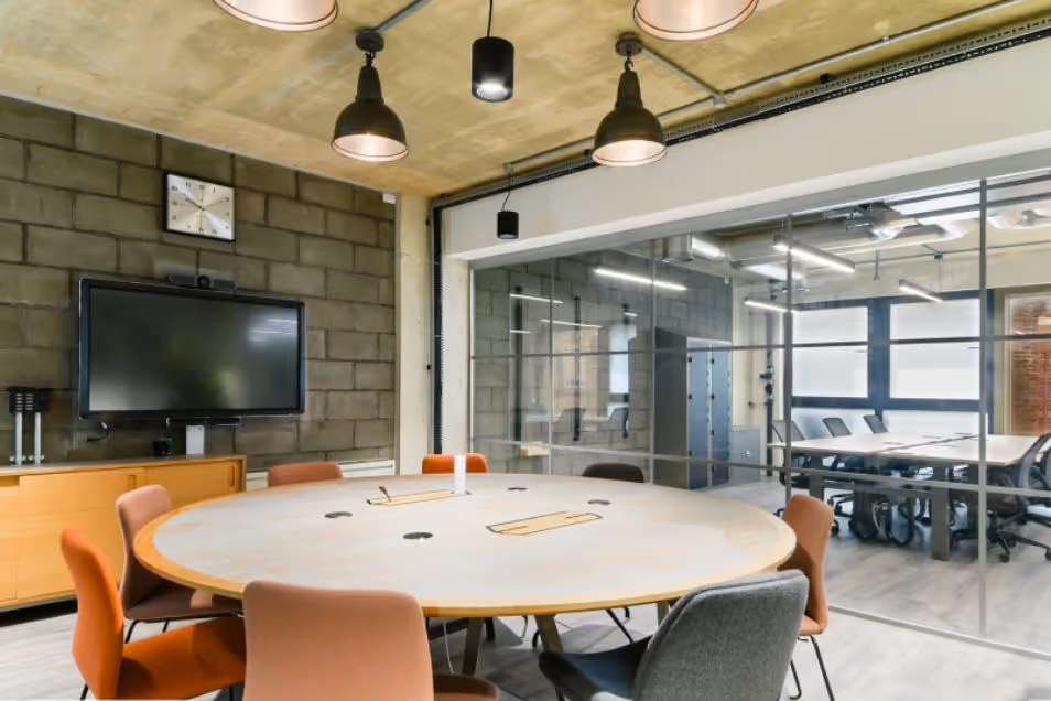 Industrial-style meeting space with a round wooden table, grey brick walls, and a glass-enclosed side office.