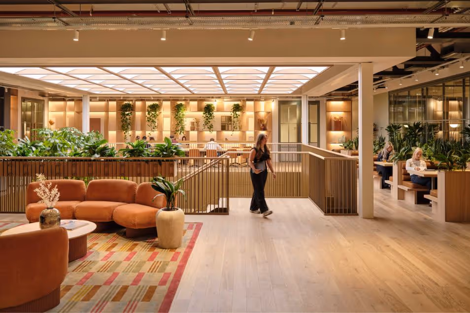 A person walks through an office lounge featuring an orange sofa, a rug, and plants under a skylight.