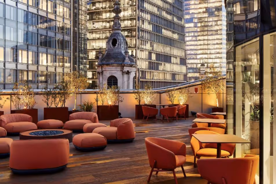 A rooftop terrace with chairs and tables overlooks city buildings and a domed structure at night.