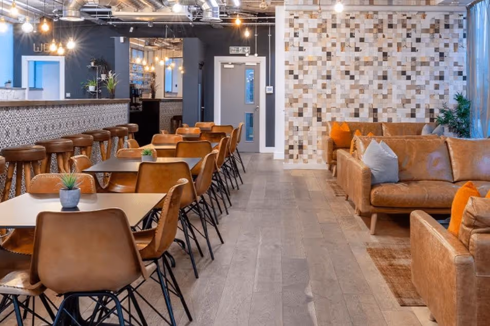 An office kitchen area with leather chairs at dining tables, a long bar, and a cosy lounge area with tan sofas.