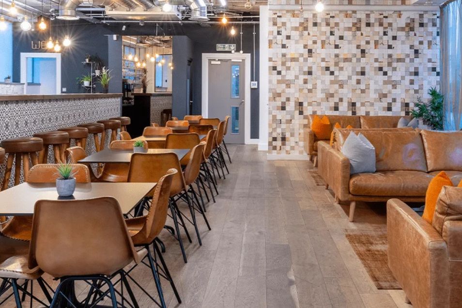 An office kitchen area with leather chairs at dining tables, a long bar, and a cosy lounge area with tan sofas.