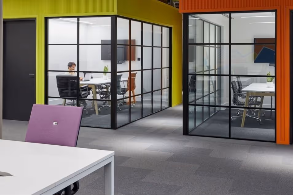 Office space with glass-partitioned meeting rooms where individuals are seated and working at white desks.