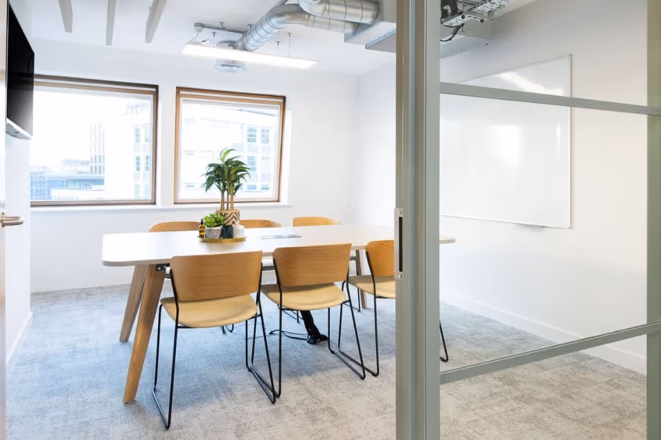 Minimalist meeting room viewed through a glass door, featuring a light wood table and matching chairs.