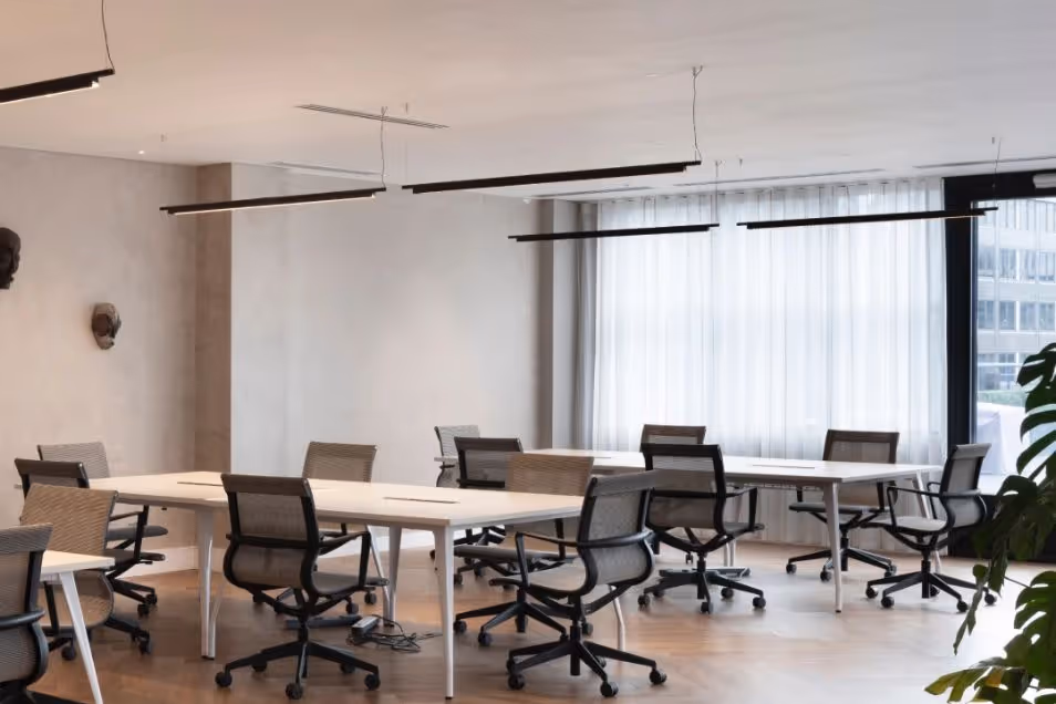 An open-plan office with white communal desks and black mesh ergonomic chairs under linear pendant lighting.