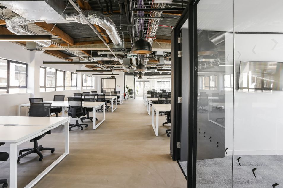Open-plan office with white desks, black mesh chairs, and glass partitions under an industrial ceiling.