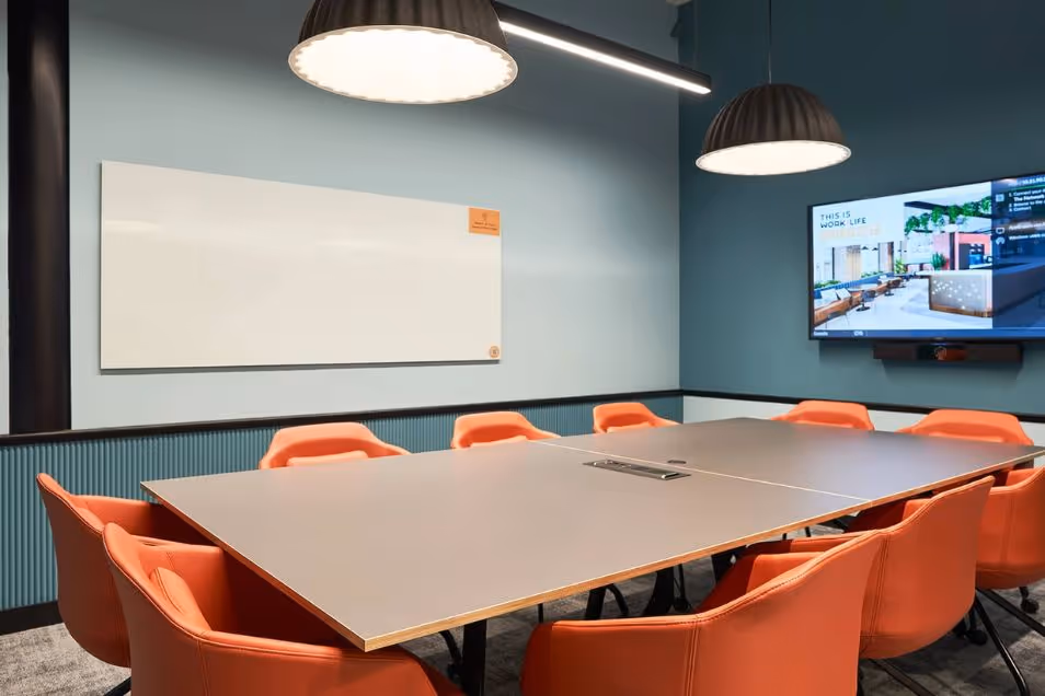 Meeting room with a large rectangular table, orange armchairs, a whiteboard, and a wall-mounted screen under pendant lighting.