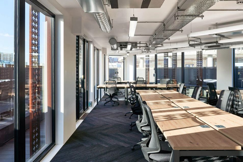 Open-plan office featuring long wooden desks, ergonomic mesh chairs, and dark chevron-patterned carpet along a row of large windows.