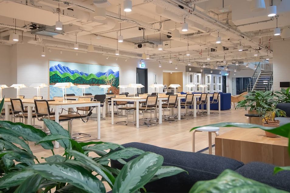 Spacious, bright coworking area with long communal tables, mushroom-style desk lamps, and a colourful mountain mural on the back wall.
