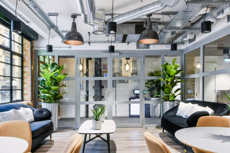 Lounge featuring plush navy and black velvet sofas, indoor plants, and a glass-paneled entrance under exposed ceiling ductwork.