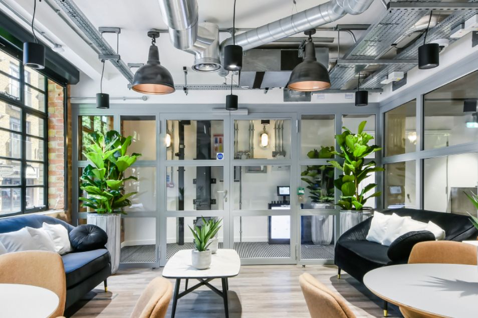 Lounge featuring plush navy and black velvet sofas, indoor plants, and a glass-paneled entrance under exposed ceiling ductwork.