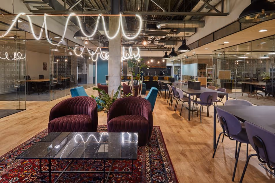 Armchairs and a coffee table on a rug in an office lounge near rows of desks and wavy ceiling lights.