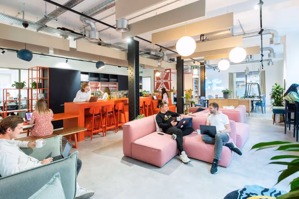 People sit on pink sofas and at a high orange table working on laptops in an open office lounge.