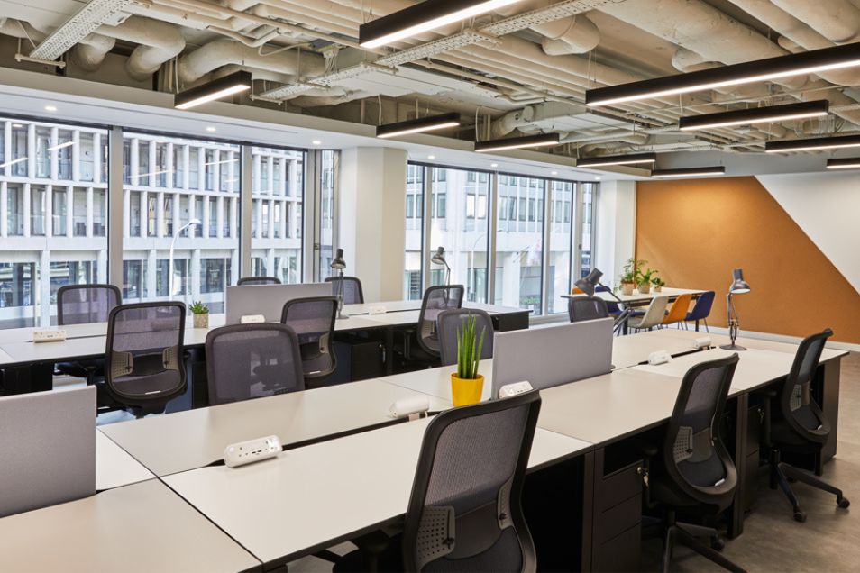 Rows of white desks with monitors and chairs in an office with large windows, a white and gold striped wall, and exposed ceiling pipes.