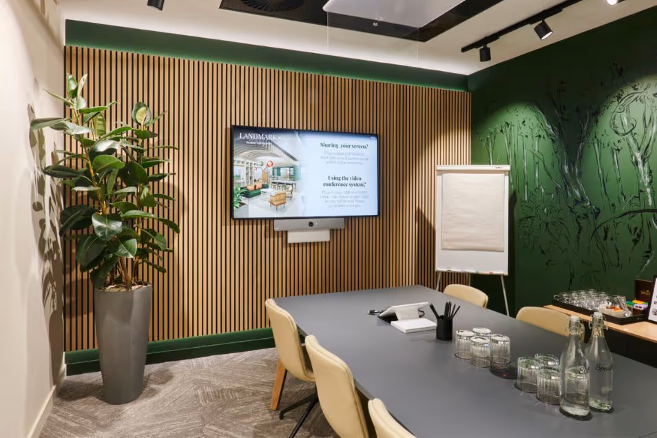 Chairs surround a table in a meeting room with a plant, a flipchart, and a screen on a wood-slat wall.