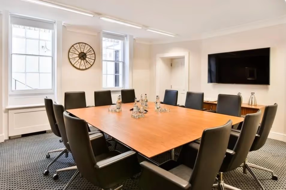 A meeting room with a wooden table and black chairs in front of a window and a clock.